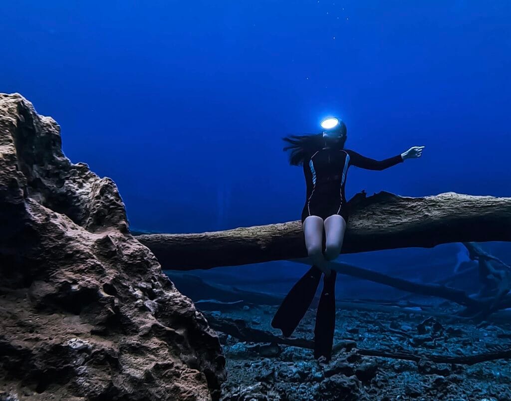 Un apnéiste (sur le Poisson des Îles) dans une combinaison de plongée noir avec une lampe frontale est assis sur un submergé journal sous l'eau, avec une grande formation rocheuse sur la gauche et un arrière-plan bleu profond.