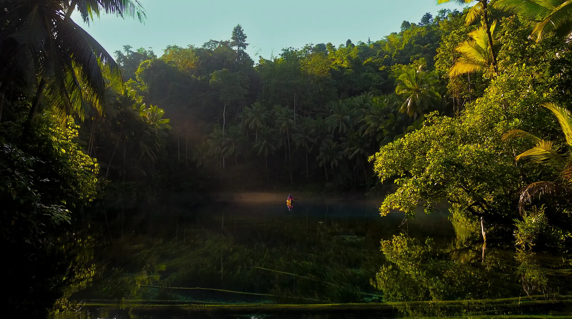 Persona remando en el lago Paisupok bajo la bóveda de selva tropical con luz filtrada entre los árboles