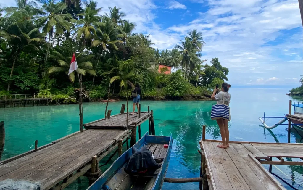 Visitors on wooden jetty at Paisu Pok Lake with turquoise water, traditional boats, coconut palms, and Indonesian flag in Banggai Islands Sulawesi